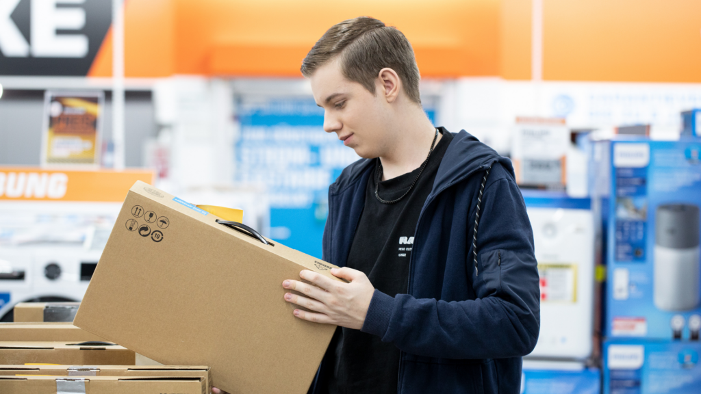 Ein junger Mann in dunkler Kleidung prüft einen großen Karton in einem Elektronikfachmarkt. Im Hintergrund sind Regale mit Haushaltsgeräten zu sehen – die Szene zeigt eine typische Arbeitssituation im Einzelhandel oder der Logistik.